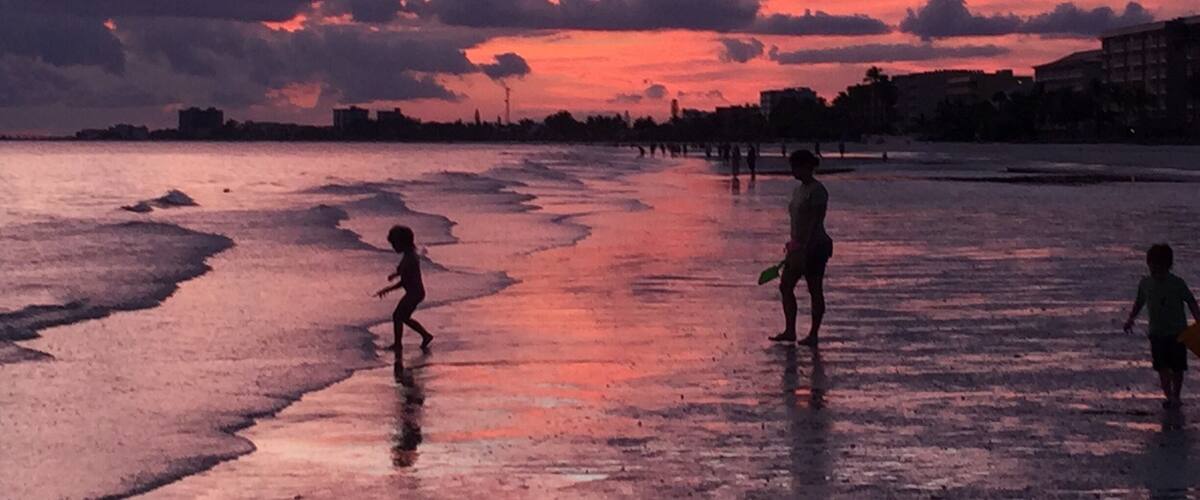 A sunset over a beach with people walking and playing in the water.