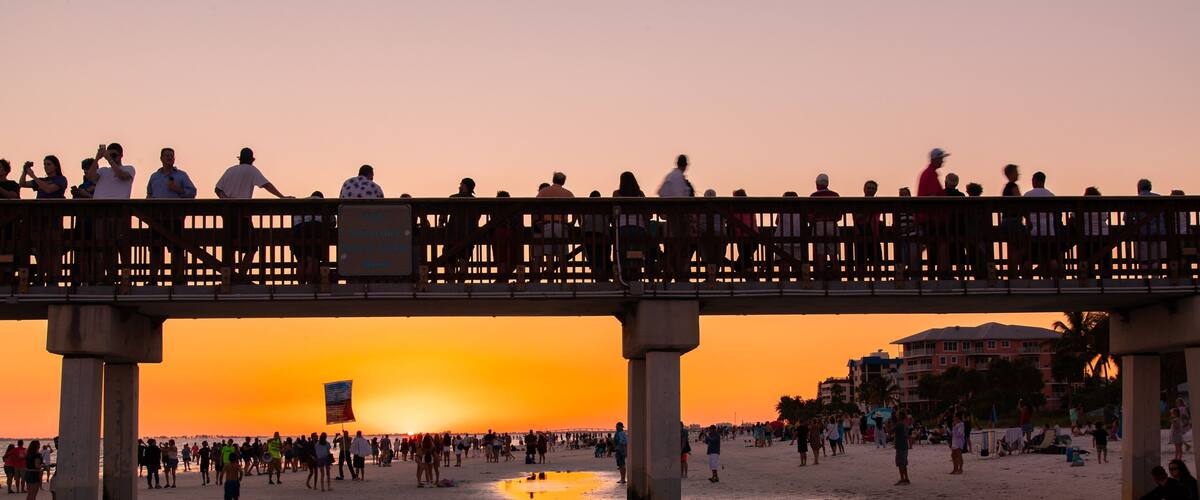 A sunset over a beach with a pier and people walking on the sand.