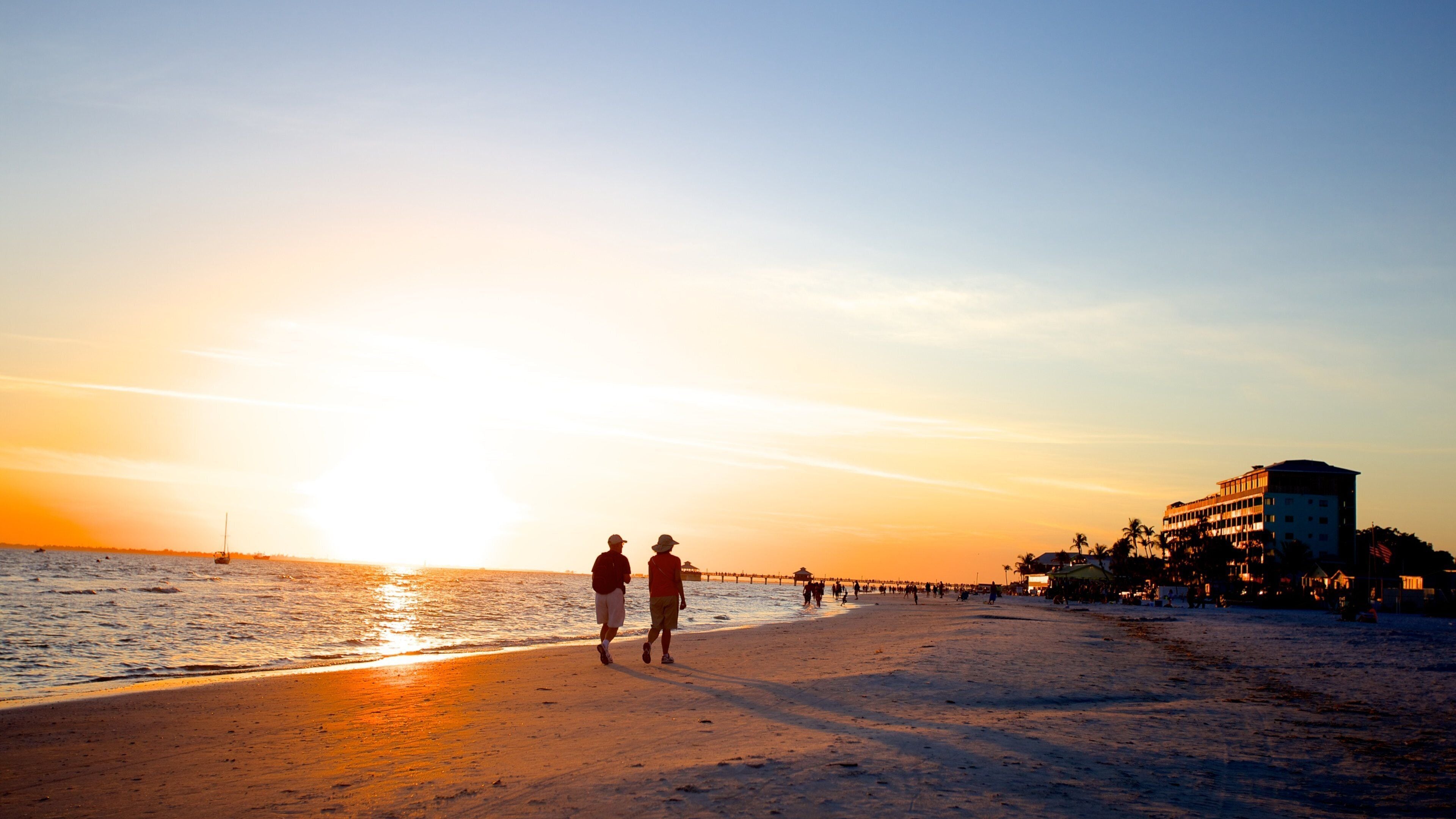 A beach at sunset with people walking along the shore, a pier extending into the water, and a building with palm trees nearby.