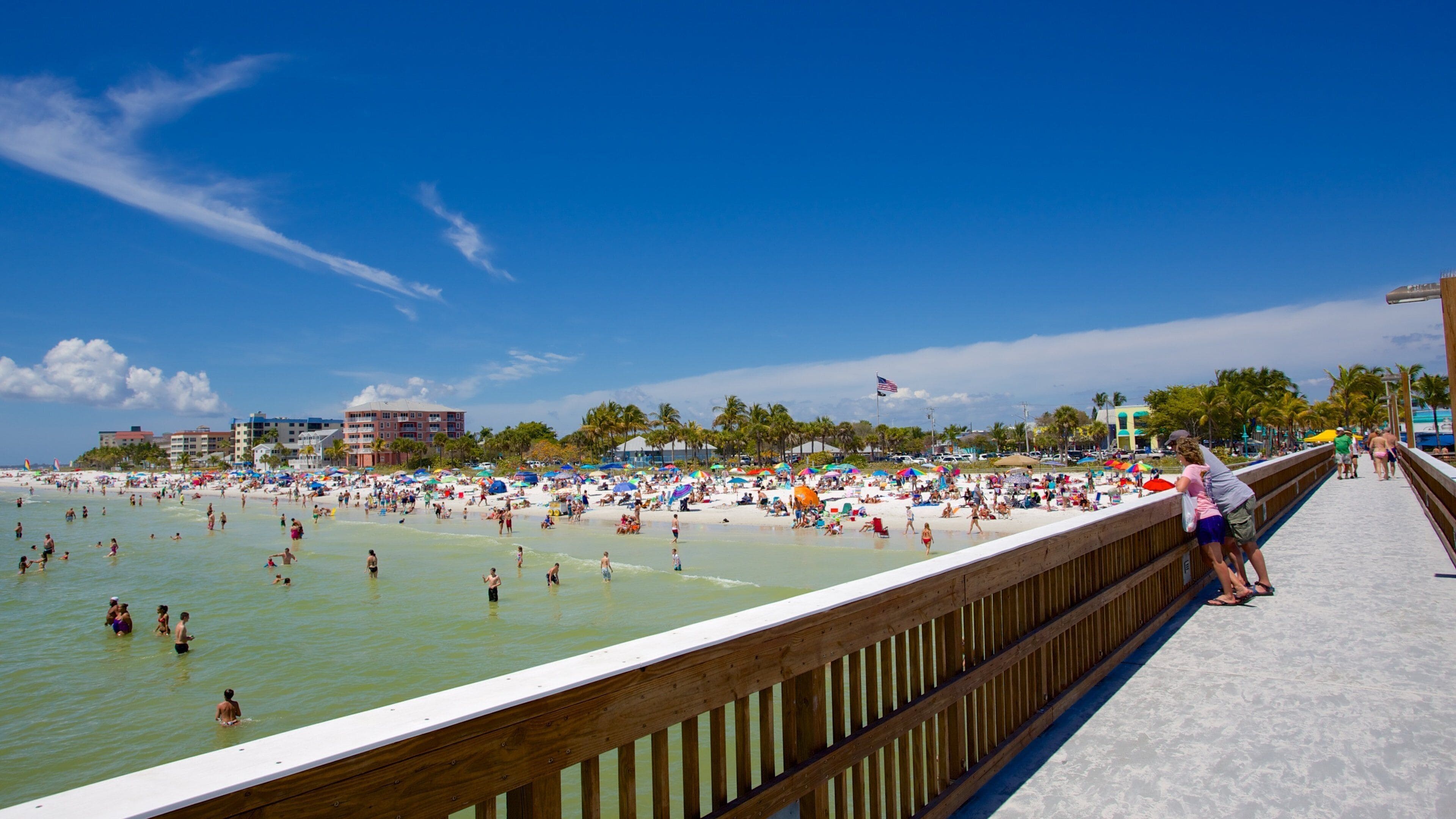 A crowded beach with people swimming and sunbathing, a wooden boardwalk, and palm trees.