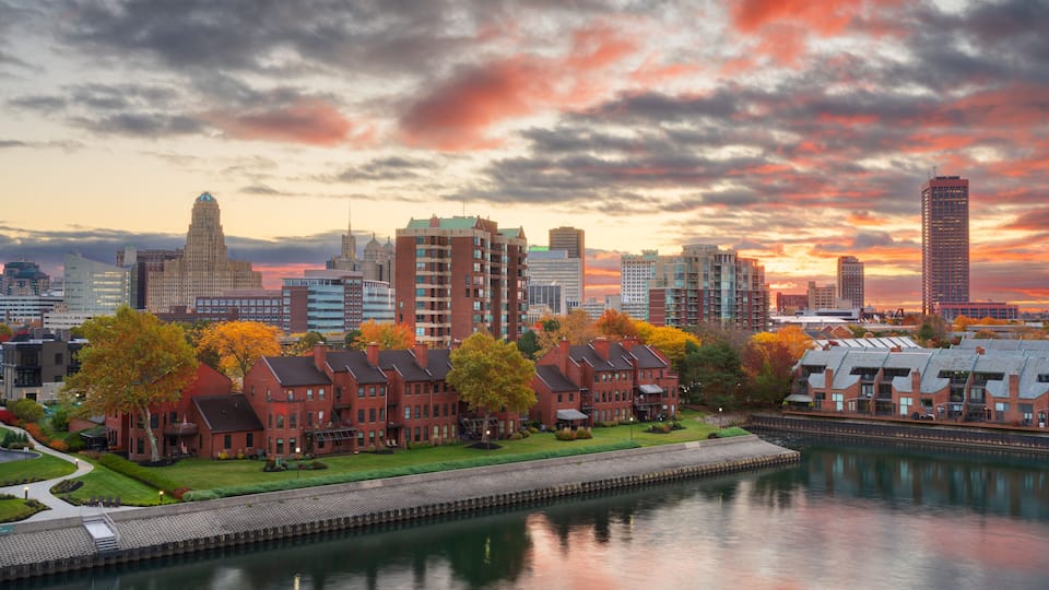 A city skyline with autumn-colored trees, a river, and a variety of buildings including a prominent skyscraper.