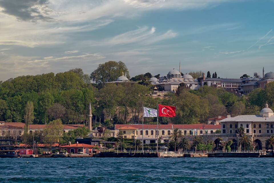 A coastal cityscape with a prominent mosque, a flag, and a historic building.