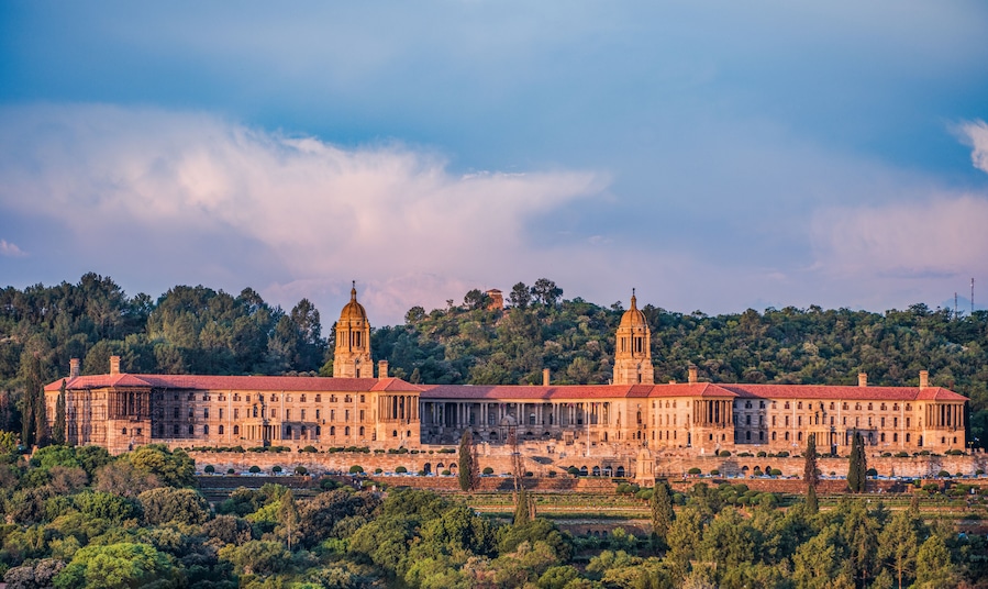 Um edifício grande e histórico com duas torres imponentes, rodeado por vegetação exuberante e um cenário de colinas.