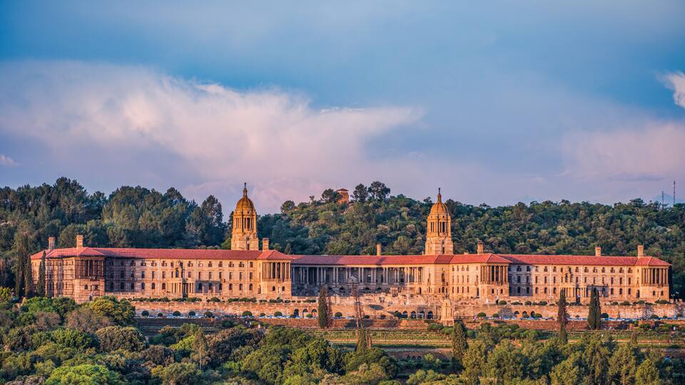 A large, historic building with two prominent towers, surrounded by lush greenery and a hilly backdrop.
