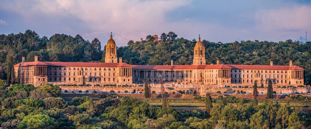 A large, historic building with two prominent towers, surrounded by lush greenery and a hilly backdrop.