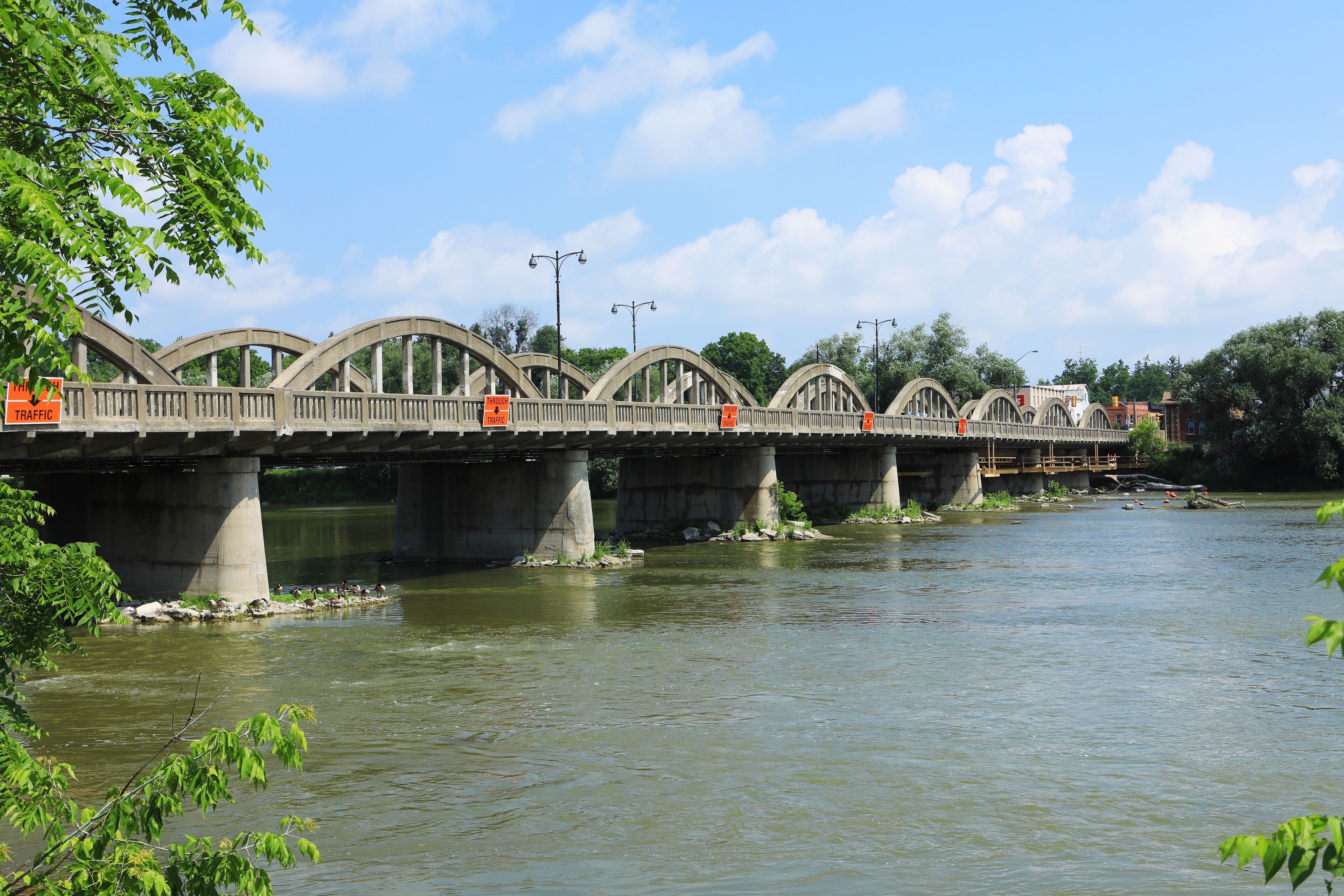 A concrete bridge with multiple arches over a river, surrounded by greenery and a clear sky.