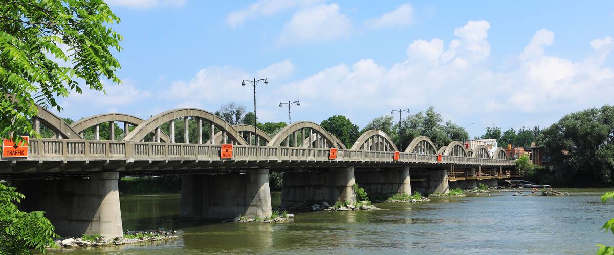A concrete bridge with multiple arches over a river, surrounded by greenery and a clear sky.