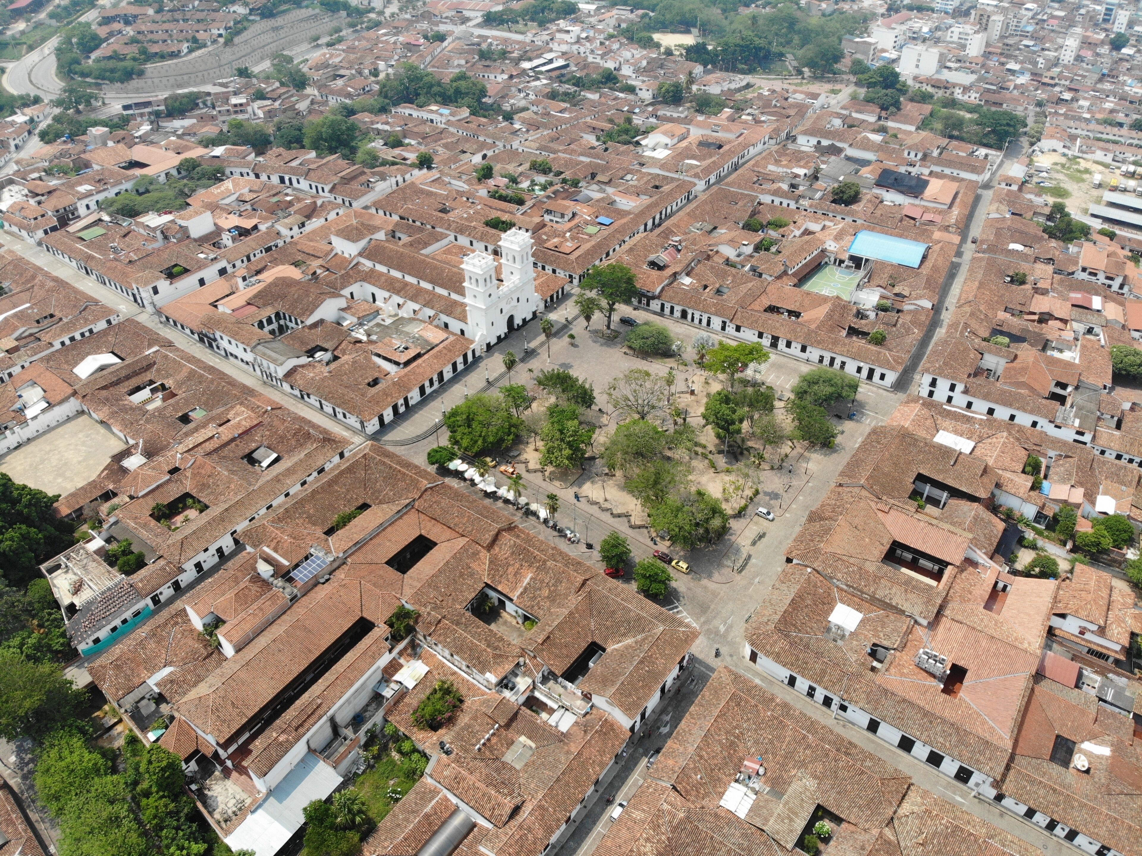 An aerial view of a densely packed urban area with terracotta-roofed buildings, a prominent white church with a bell tower, and a central open square surrounded by greenery.