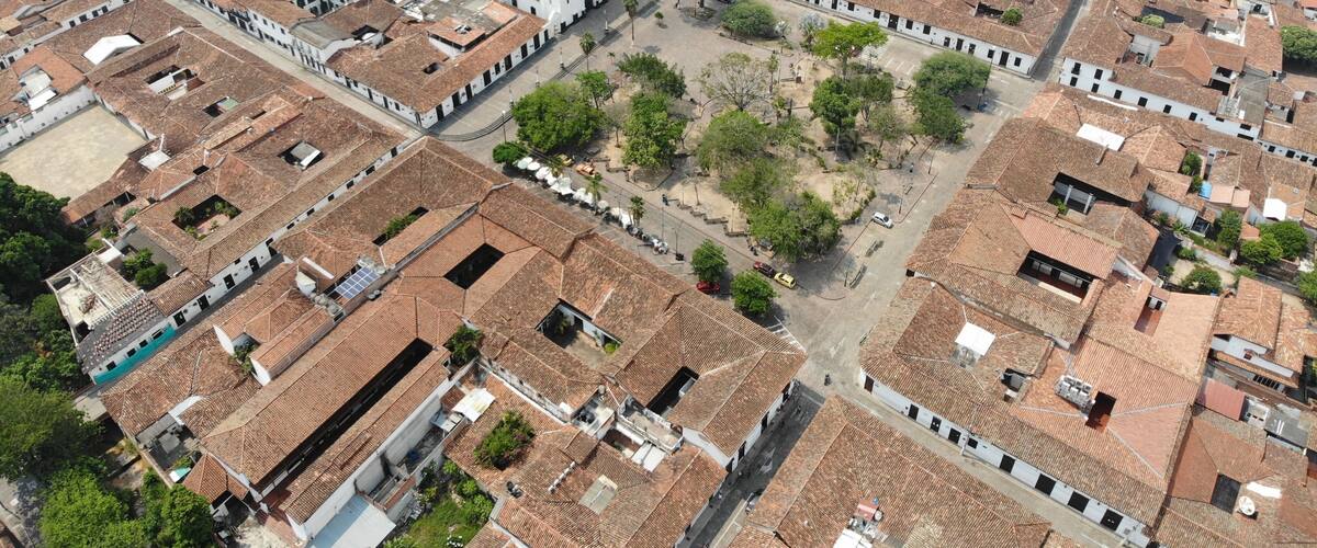 An aerial view of a densely packed urban area with terracotta-roofed buildings, a prominent white church with a bell tower, and a central open square surrounded by greenery.