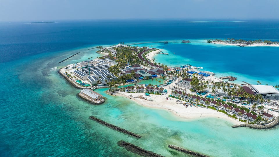 An aerial view of a tropical island resort with clear turquoise waters, a sandy beach, and a variety of buildings including a large hotel complex.