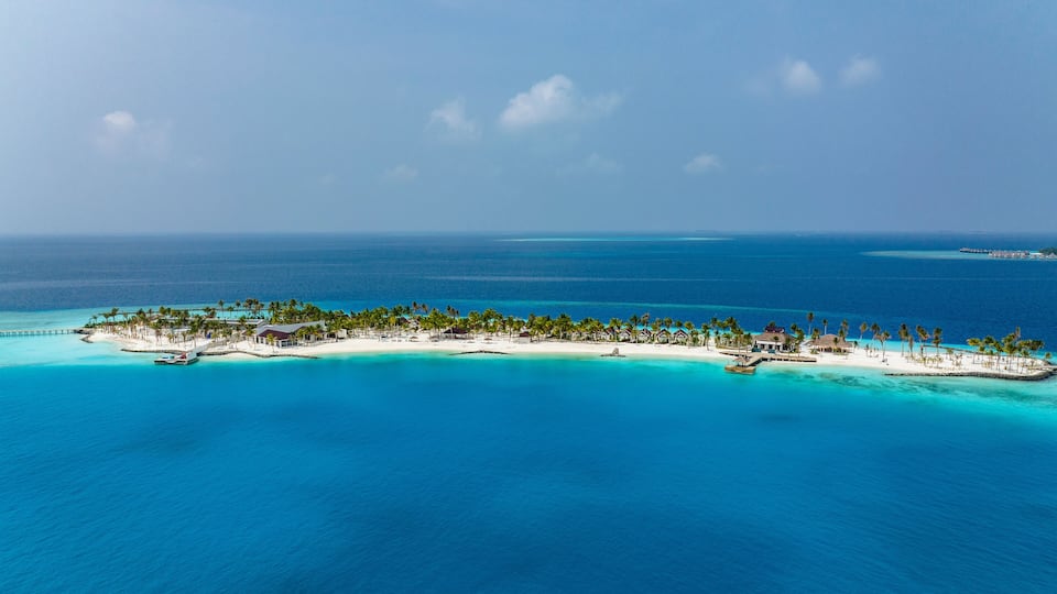 An aerial view of a tropical island with clear turquoise waters, white sandy beaches, and a row of palm trees.