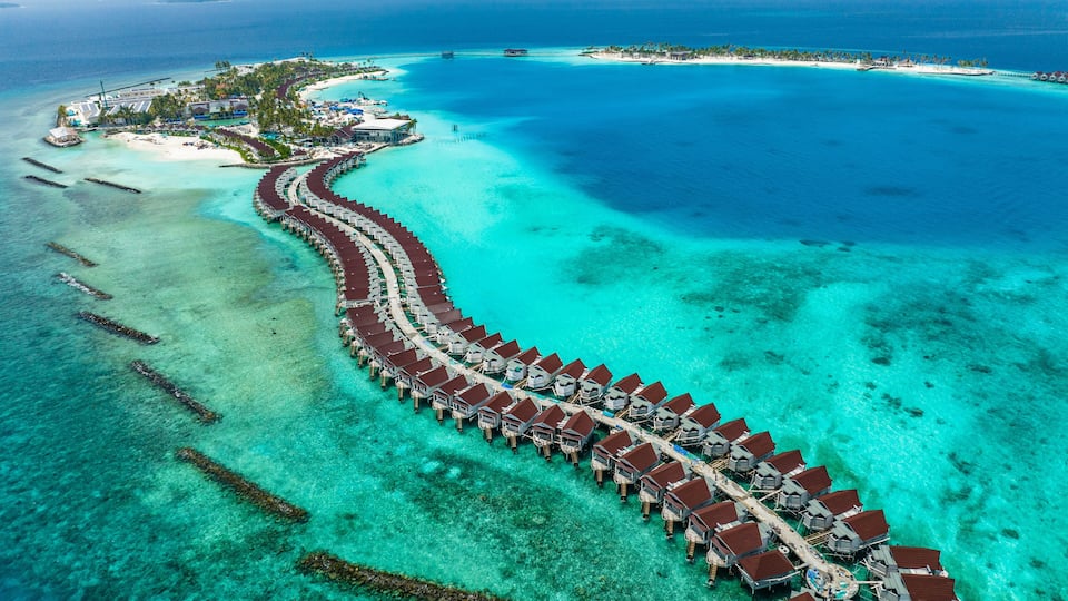 An aerial view of overwater bungalows connected by a pier, surrounded by turquoise waters and a small island with buildings.