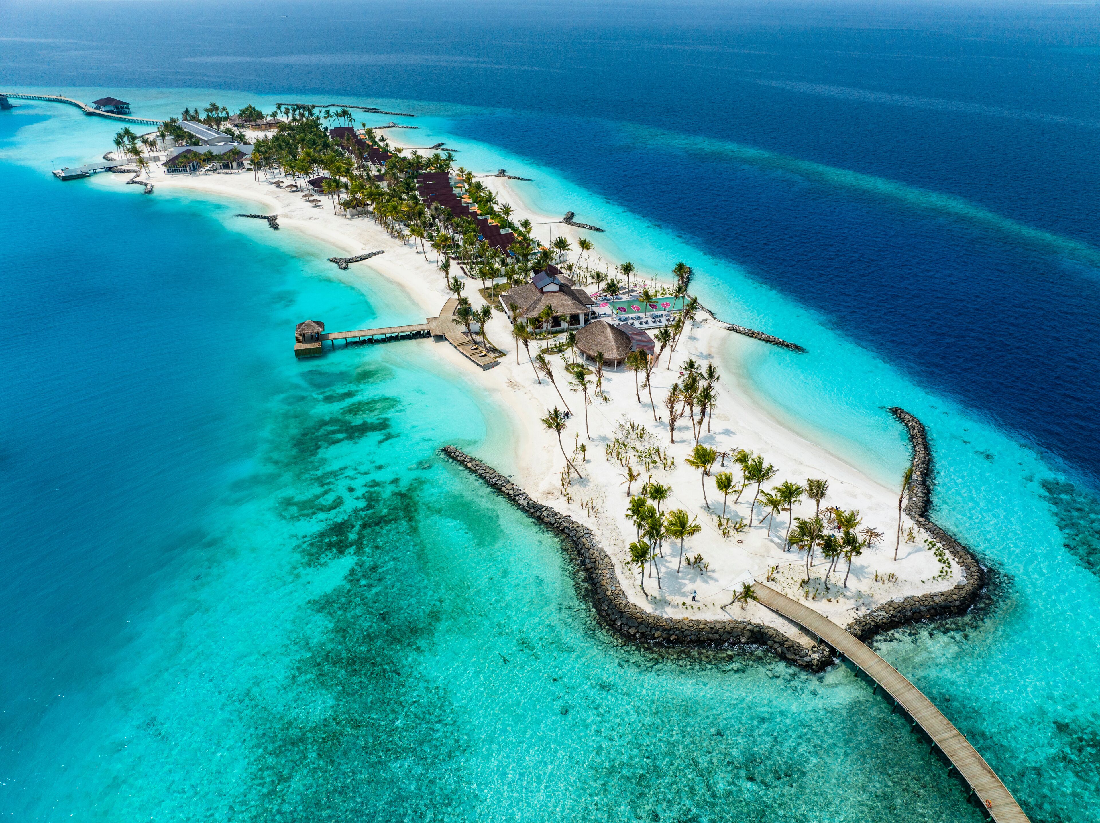 An aerial view of a tropical island with a white sandy beach, clear turquoise waters, and a wooden pier connecting to the mainland.