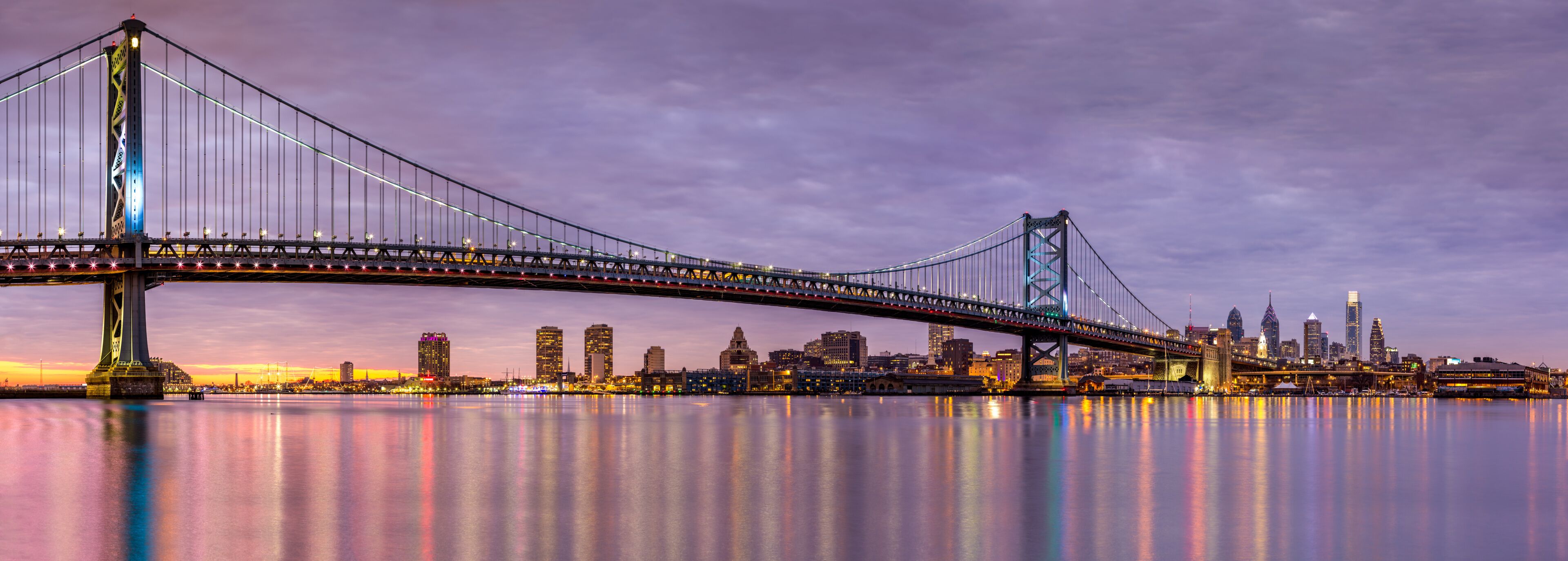 A large suspension bridge spanning a body of water with a city skyline in the background.
