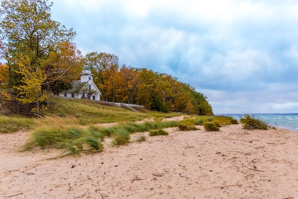 En strand med et fyrtårn, efterårstræer og en overskyet himmel.