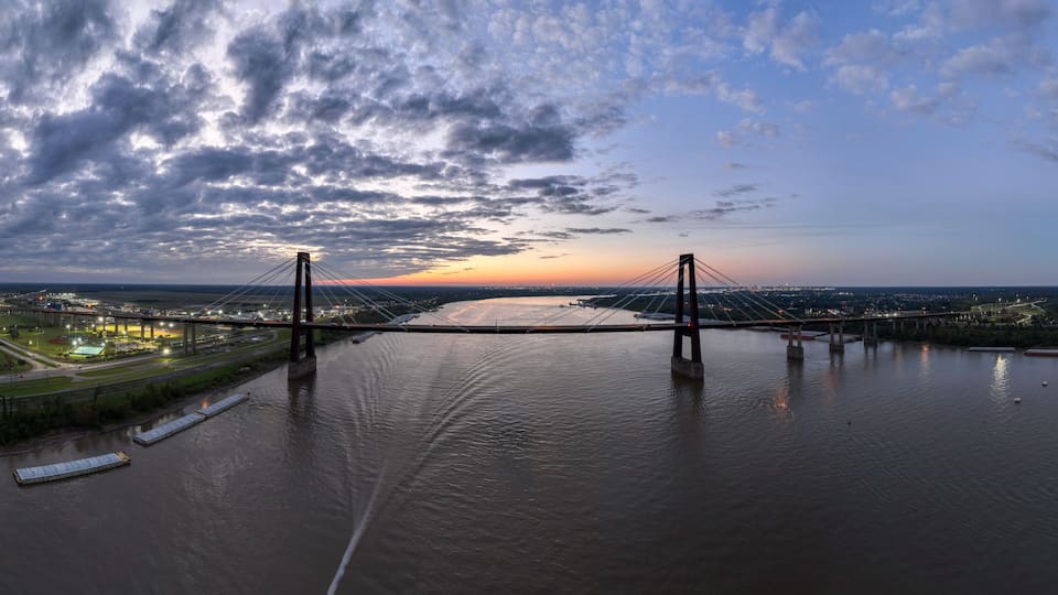A large suspension bridge spanning a wide river, surrounded by urban areas and greenery.