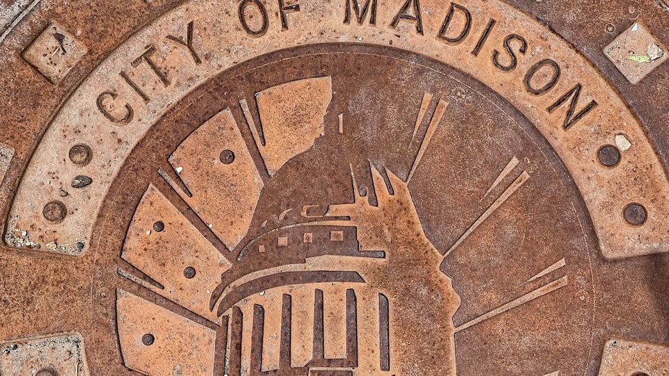 A weathered metal plaque with the inscription "CITY OF MADISON" and a depiction of a building, likely a government or historical structure.