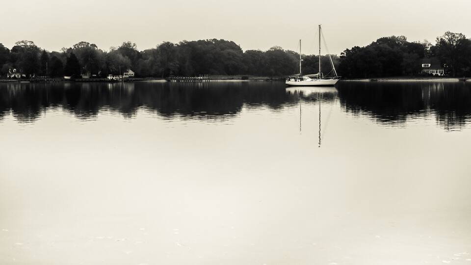A calm lake with a sailboat, trees, and a house on the shore.