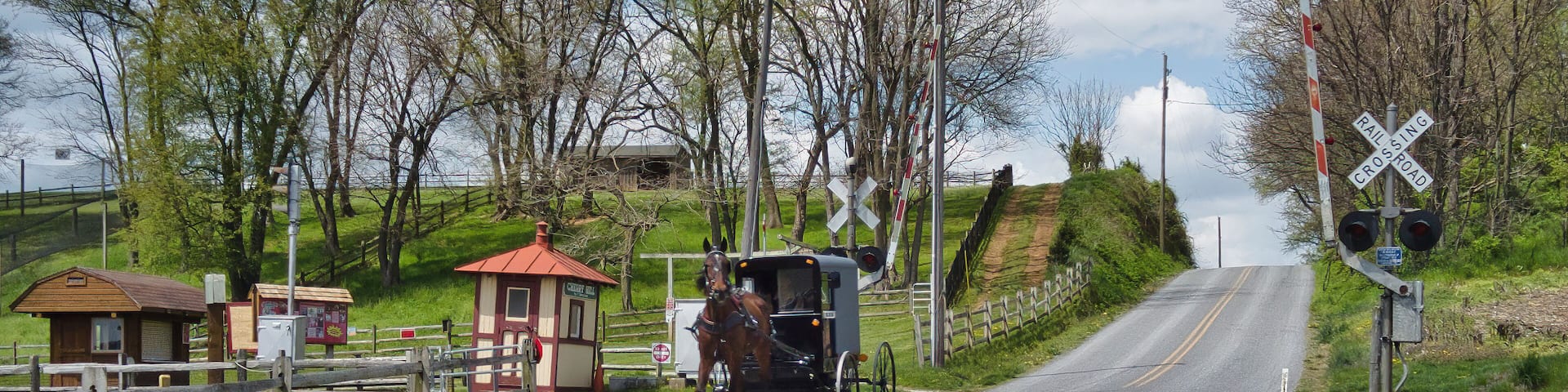 A rural road with a horse-drawn carriage, a small wooden building, and a railroad crossing.