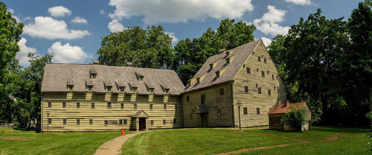 A historic wooden building with a steeply pitched roof, surrounded by lush green trees and a clear blue sky.