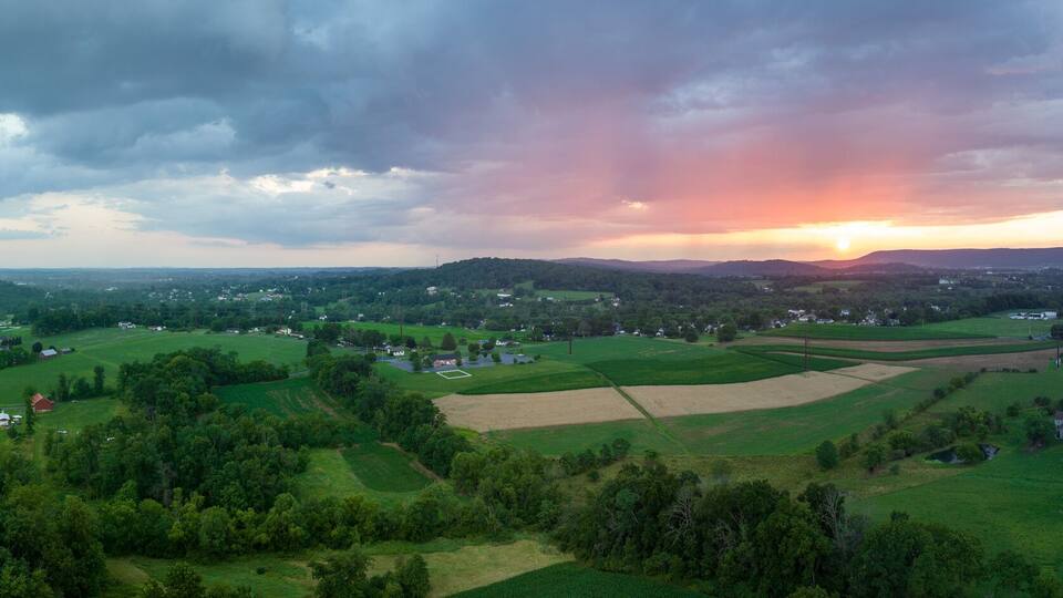A rural landscape with fields, scattered houses, and a sunset over rolling hills.