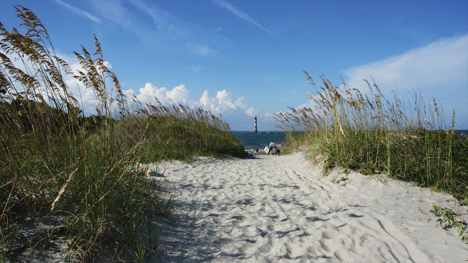 A sandy path leading to a lighthouse, surrounded by tall grasses and a clear blue sky.