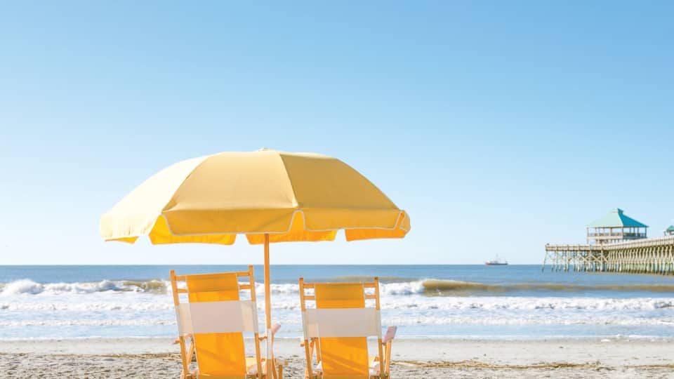 A beach with two yellow beach chairs and a large yellow umbrella. In the background, there is a pier with a blue roof and a ship in the water.