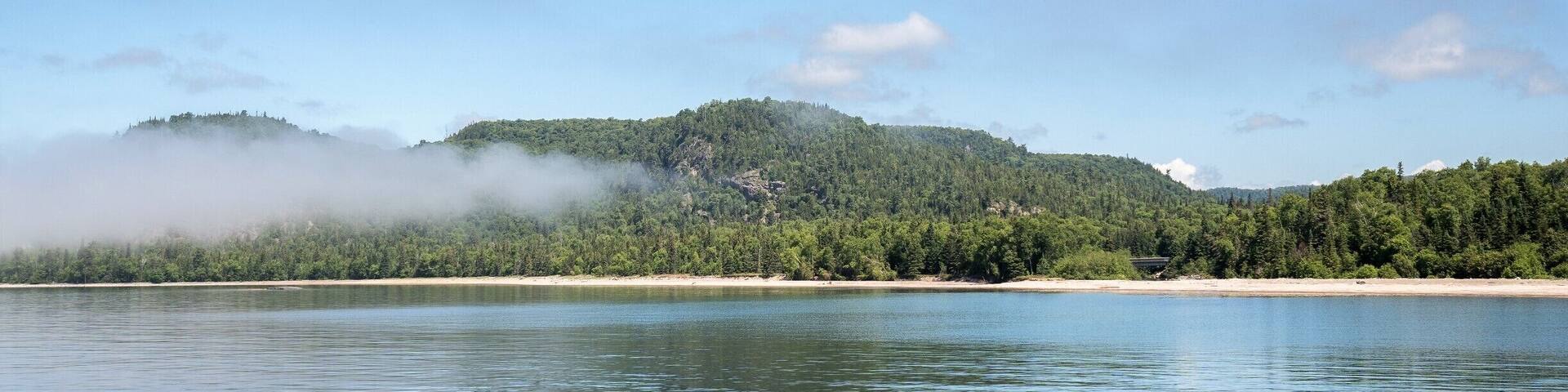 A calm lake with a forested hill in the background and a clear blue sky.