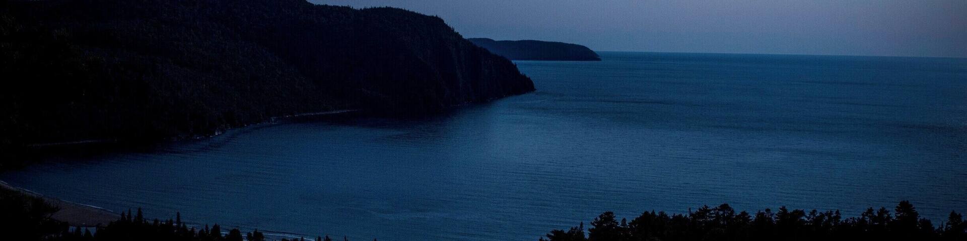 Un paysage côtier au crépuscule, avec une mer calme, une île lointaine et une côte bordée d’une forêt.