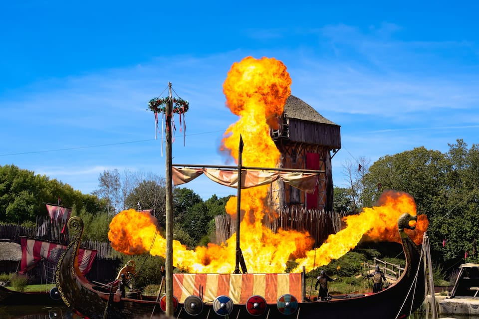 A Viking ship engulfed in flames with a wooden structure in the background.