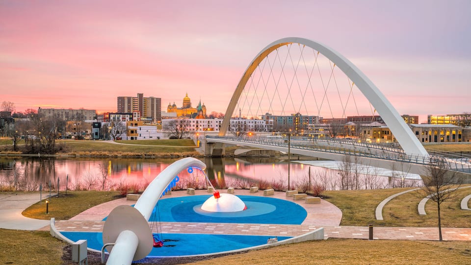 A park with a large white arch bridge, a domed building, and a calm lake reflecting the sunset.