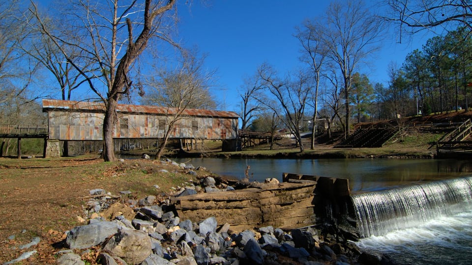 A river with a waterfall, a rusted building, and leafless trees.