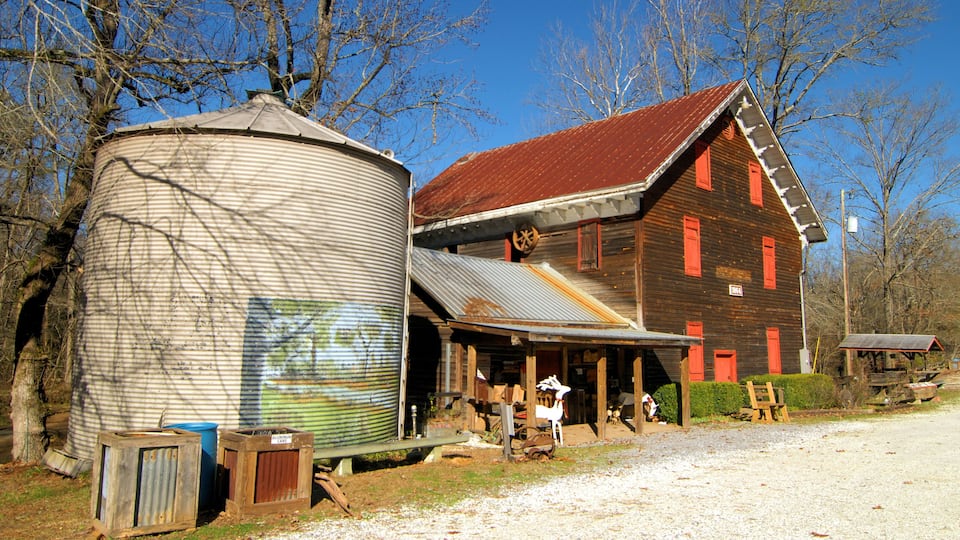 A rustic barn with a red roof and wooden siding, a large cylindrical grain silo, and a small shed in a rural setting.