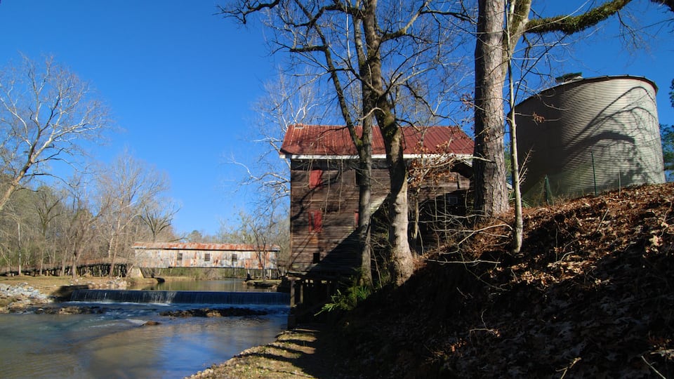 A rustic wooden building with a red roof near a calm river, surrounded by leafless trees and a metal water tank.