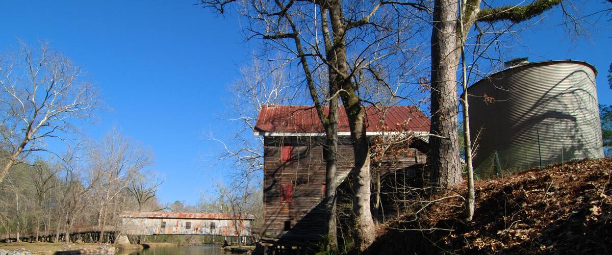 A rustic wooden building with a red roof near a calm river, surrounded by leafless trees and a metal water tank.