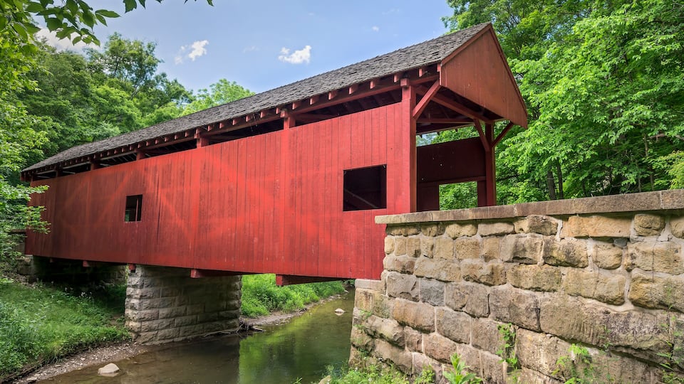 A red covered bridge spanning a calm river, surrounded by lush greenery and stone walls.