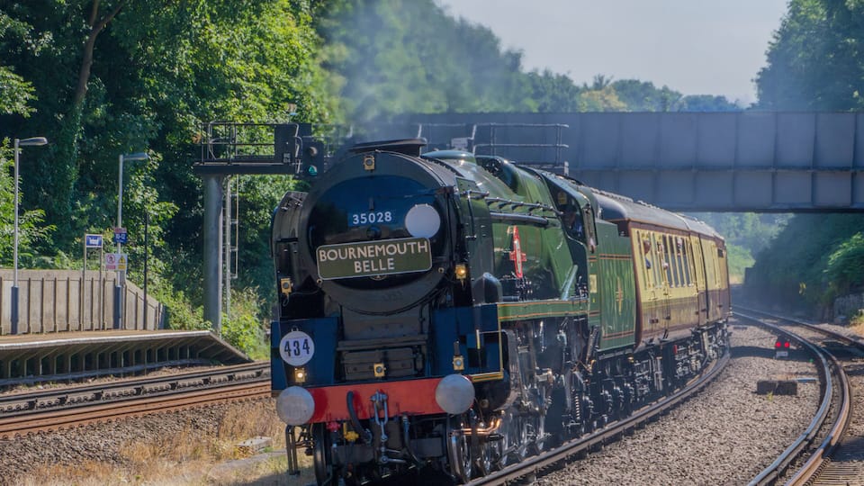 A train with the number 35028 labeled 'Bournemouth Belle' traveling on tracks with a bridge overhead.