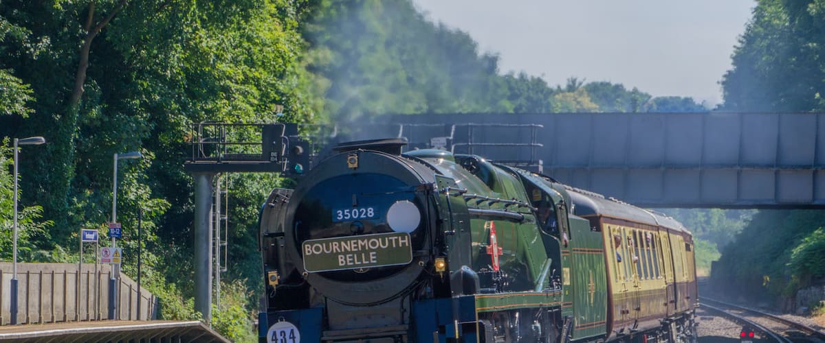 A train with the number 35028 labeled 'Bournemouth Belle' traveling on tracks with a bridge overhead.