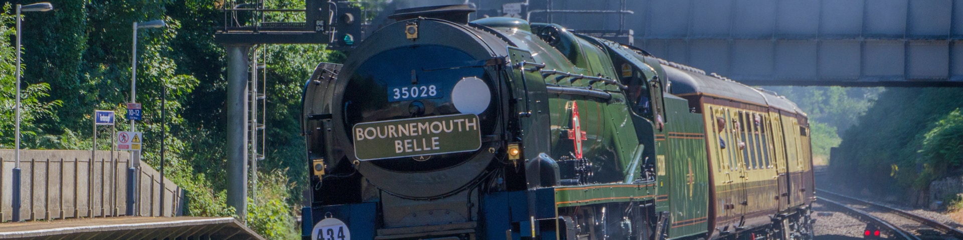 A train with the number 35028 labeled 'Bournemouth Belle' traveling on tracks with a bridge overhead.