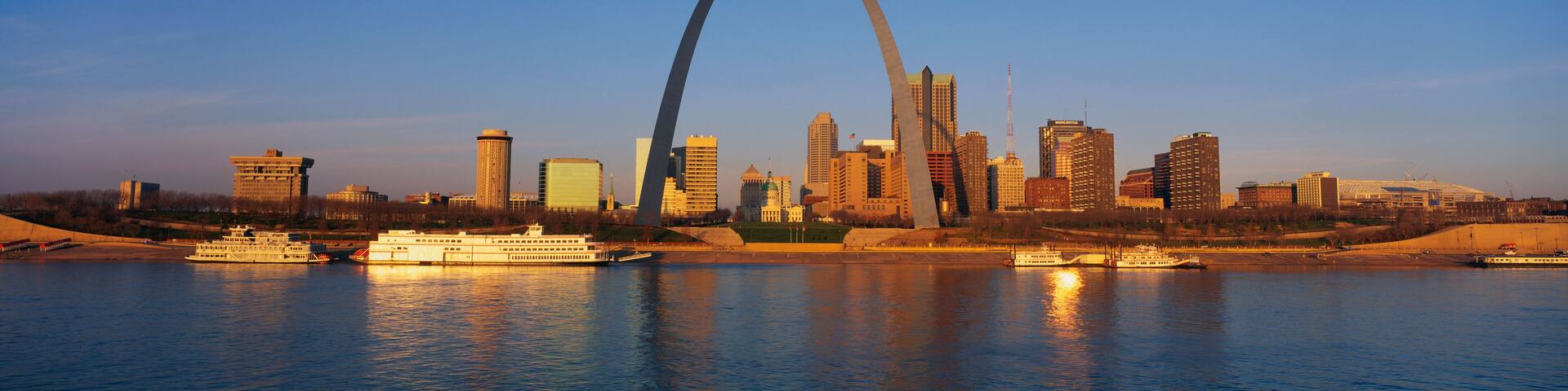 This is the skyline at sunrise. It is situated along the Mississippi River. There are riverboats on the water with the St. Louis Arch in clear view.