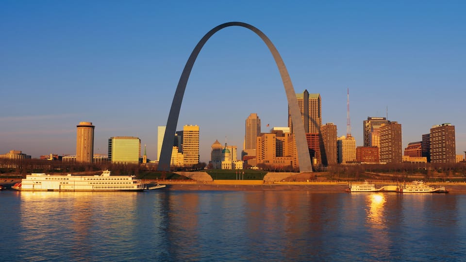 A city skyline with a prominent arch bridge over a river, featuring several high-rise buildings and boats on the water.