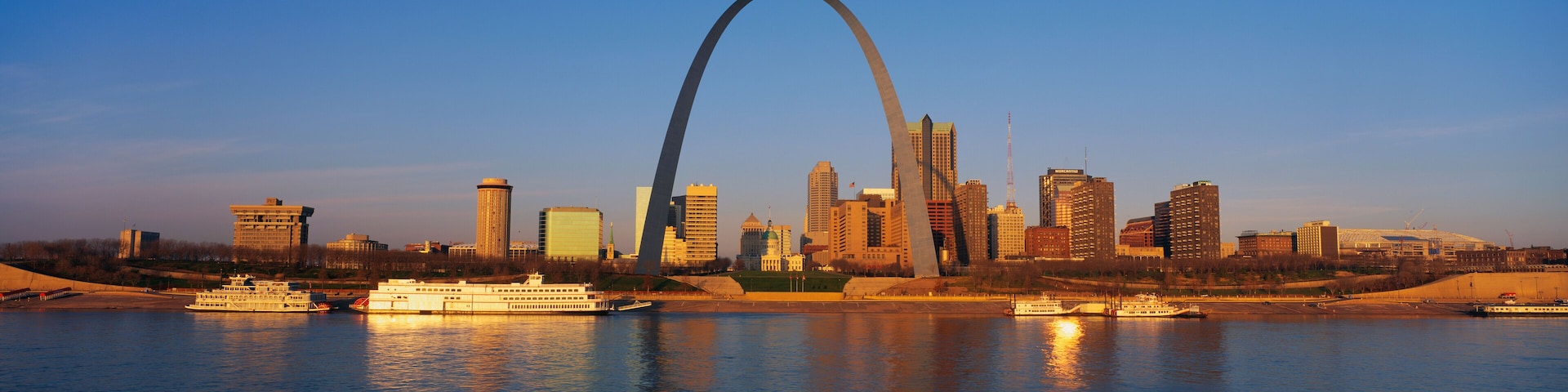 This is the skyline at sunrise. It is situated along the Mississippi River. There are riverboats on the water with the St. Louis Arch in clear view.