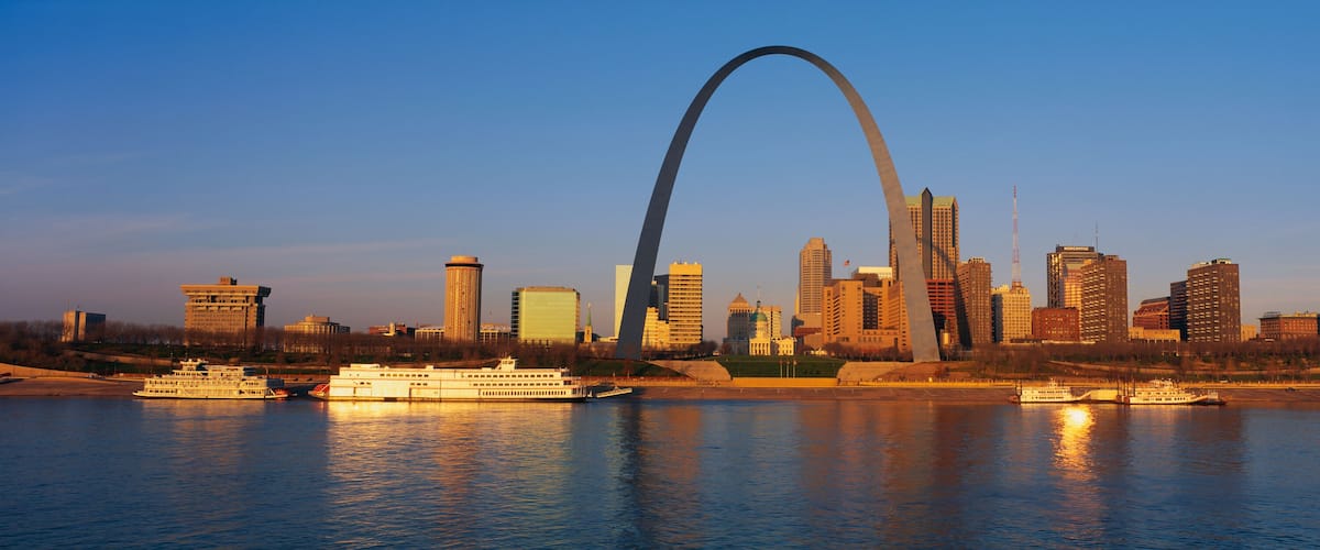 A city skyline with a prominent arch bridge over a river, featuring several high-rise buildings and boats on the water.