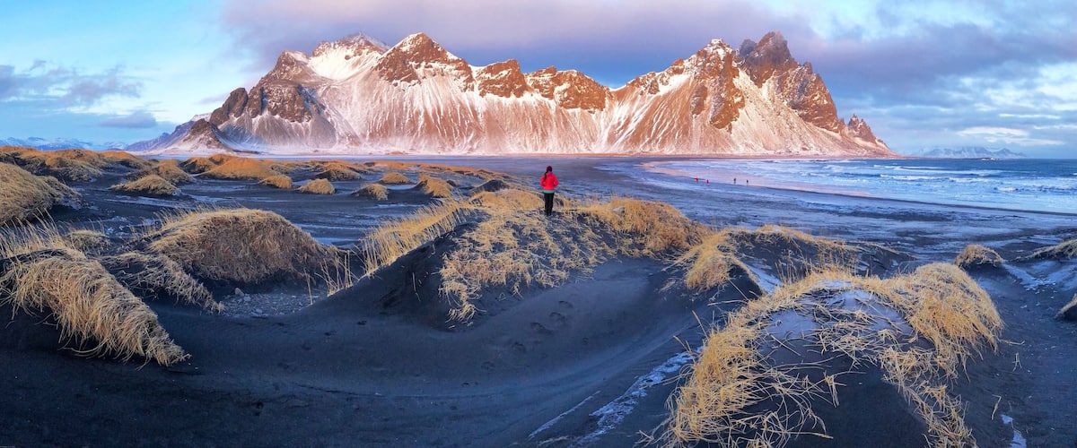 A person standing on a beach with black sand and grassy dunes, with a mountain range in the background.