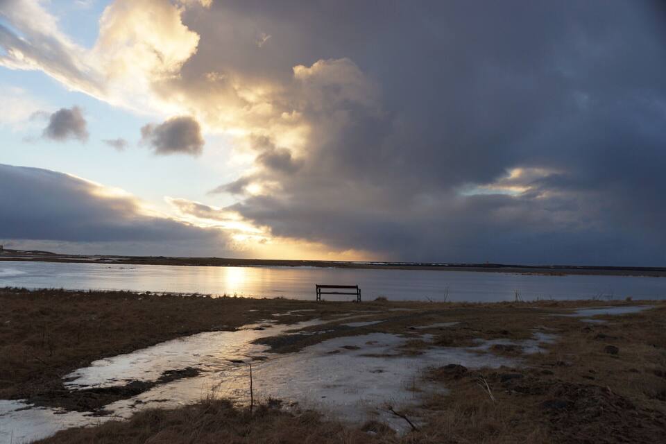 A bench overlooking a coastal landscape with a sunset and stormy sky.