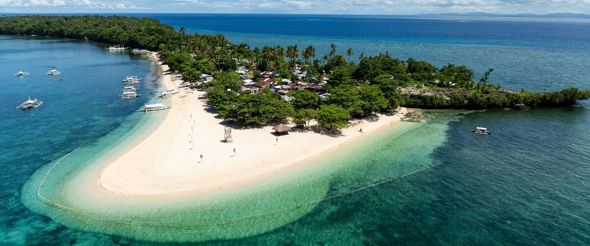 Eine Vogelperspektive einer tropischen Insel mit einem weißen Sandstrand, klarem türkisblauem Wasser und vereinzelten Booten.