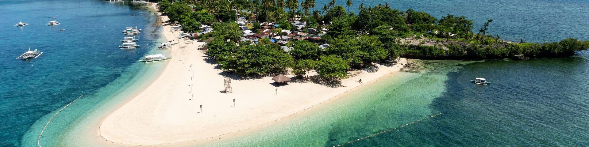 White sand beach meets turquoise water beside trees and houses. Tulang Diot. Camotes, Philippines.