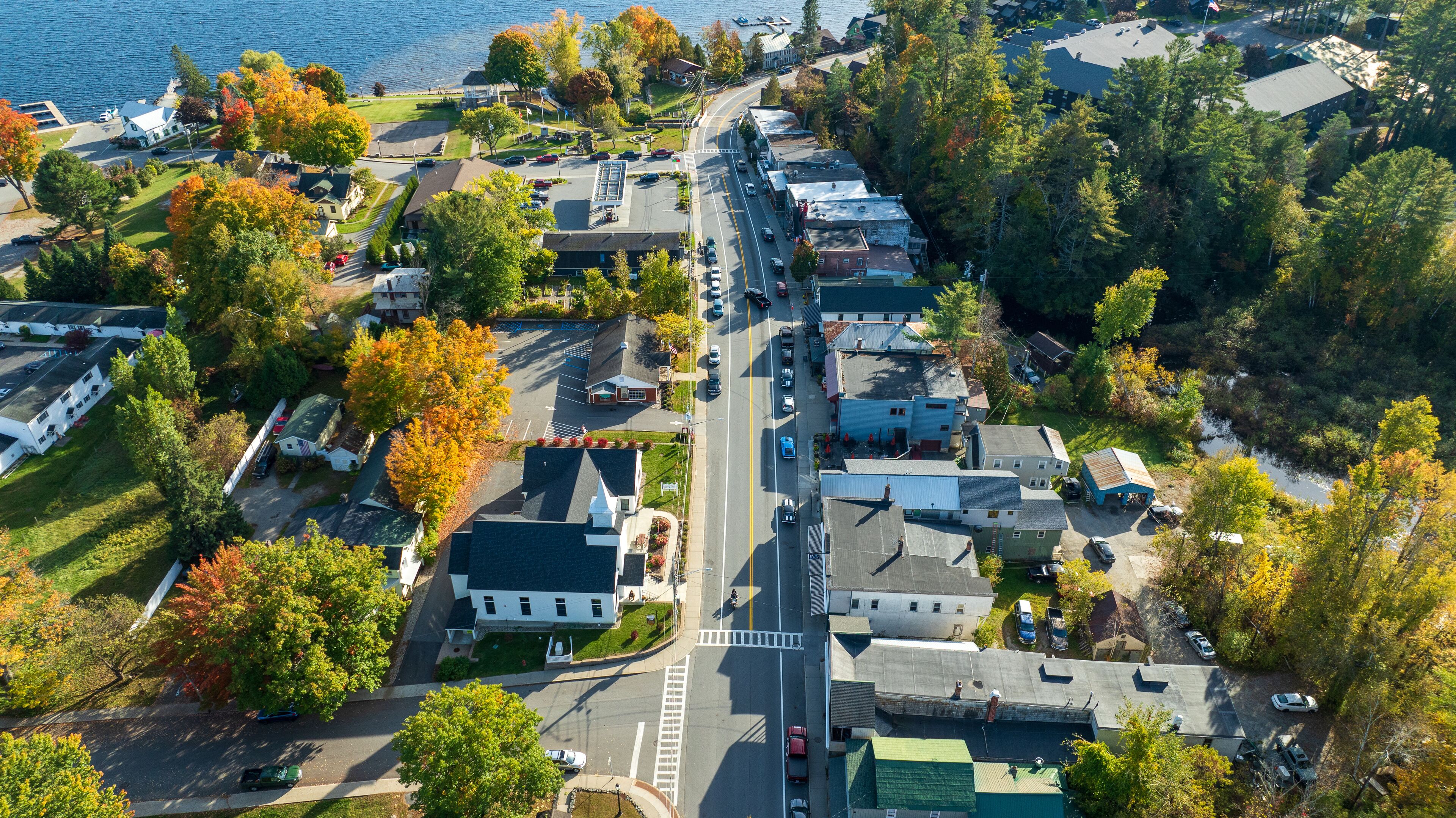 Aerial view of Schroon Lake, New York, showcasing the calm lake, fall foliage, and small town structures nestled along the shoreline under a clear sky.