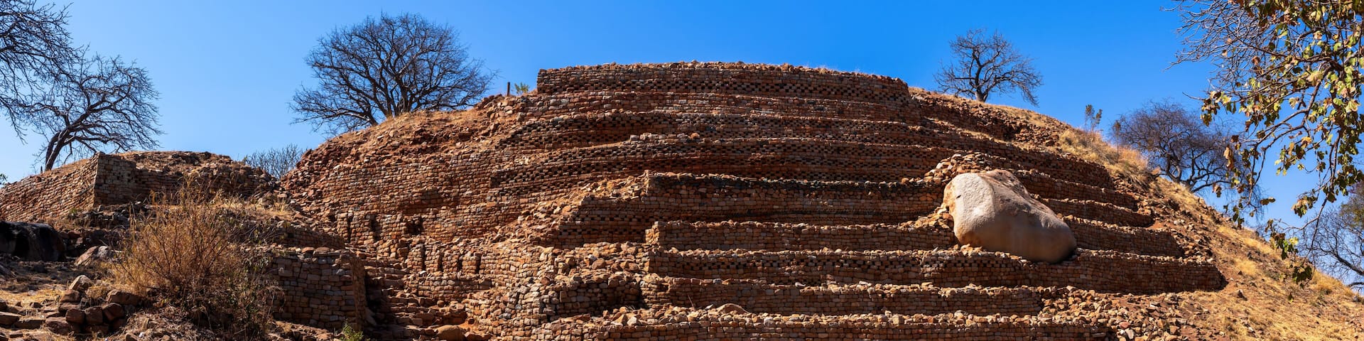 Ancient brick ruins with a large stone slab on top, surrounded by dry, rocky terrain and sparse vegetation.