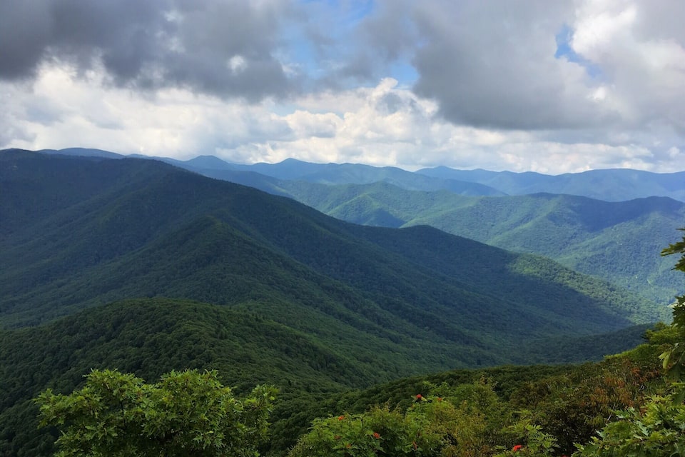 A mountainous landscape with dense green forests and distant mountain ranges under a partly cloudy sky.